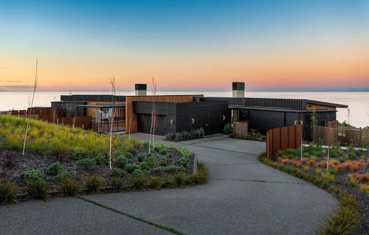 Black and cedar house on the hill above the bay at sunset, with a curving driveway and landscaped front garden