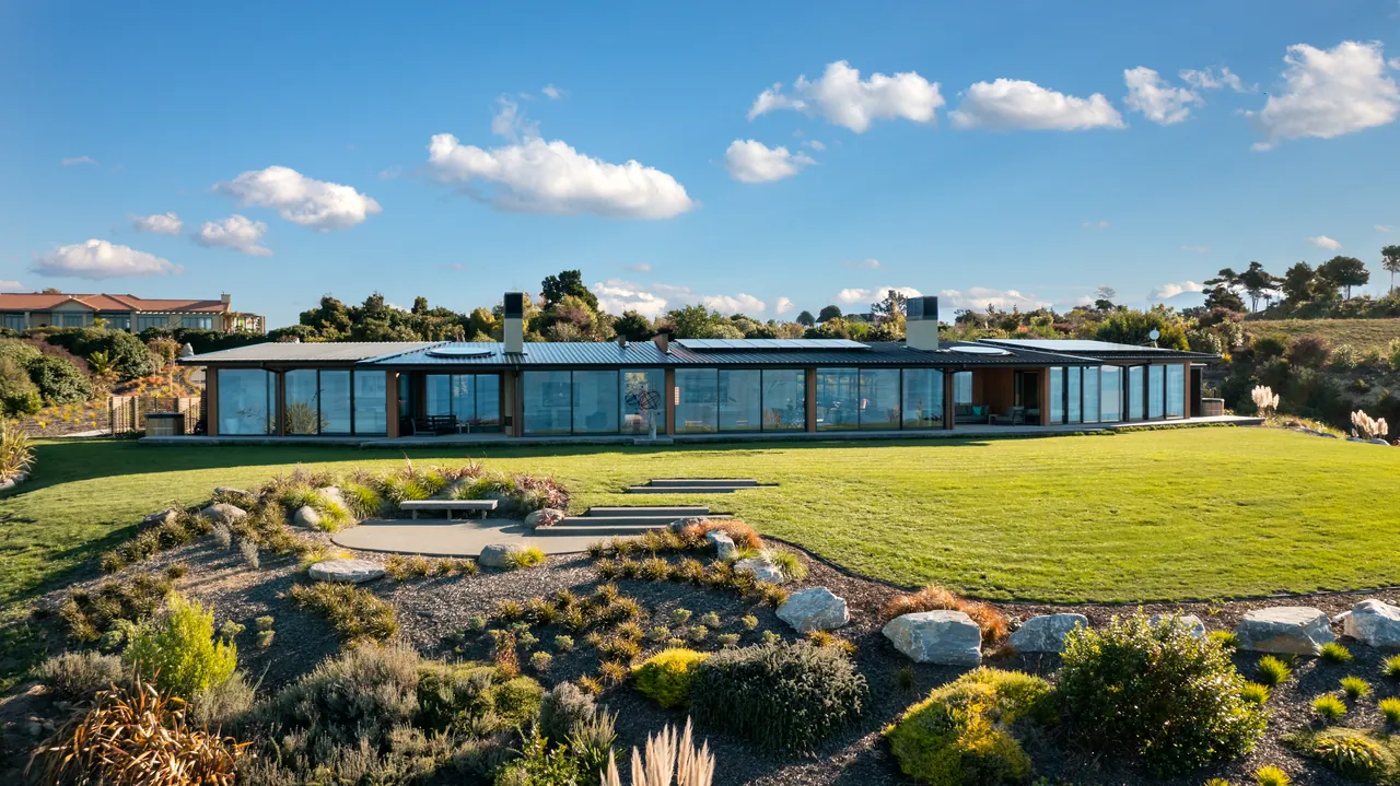 Modern glass-walled house with a metal roof set among native planting, rocks and lawn under a blue sky